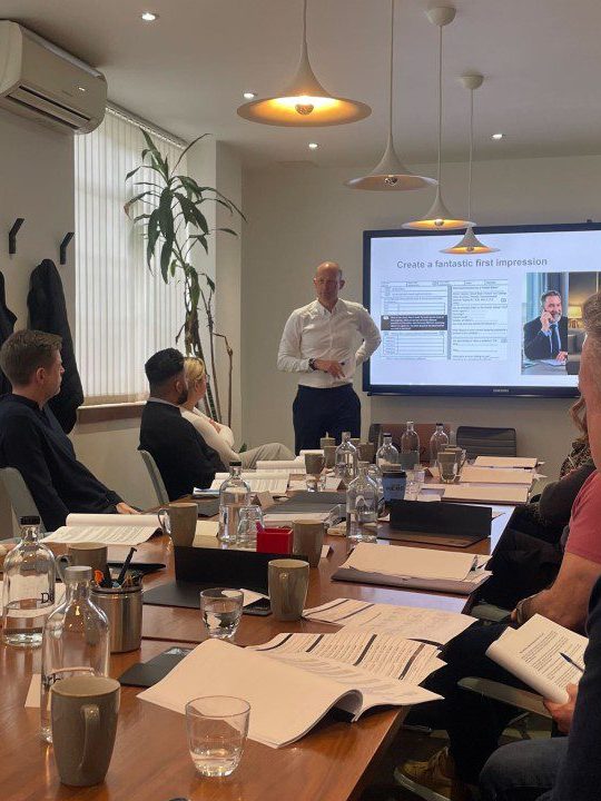 A group of people sitting around a table in a meeting room, with one individual standing and presenting information on a screen in the background.